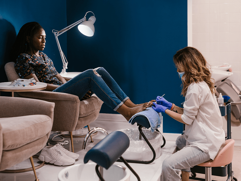 A female nail technician curing feet nail of black girl at salon, sitting on couch.